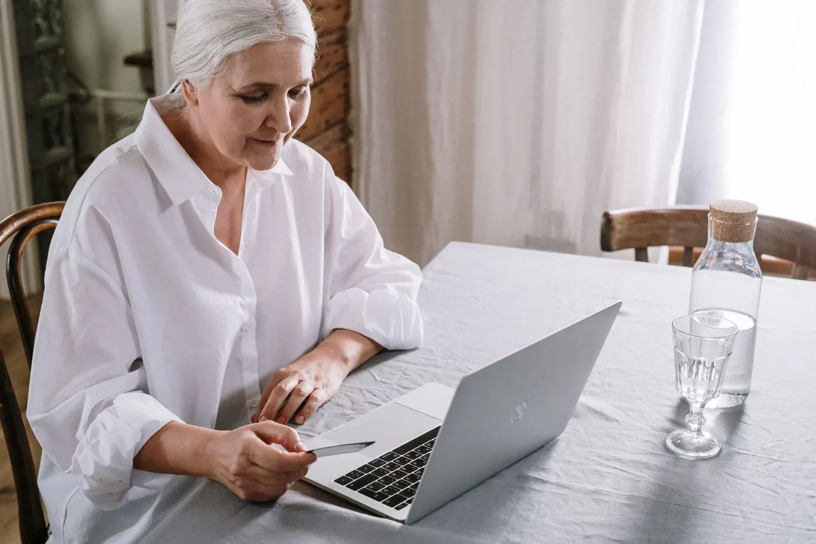Mujer mayor comprando en línea con su laptop y tarjeta en una mesa con luz natural